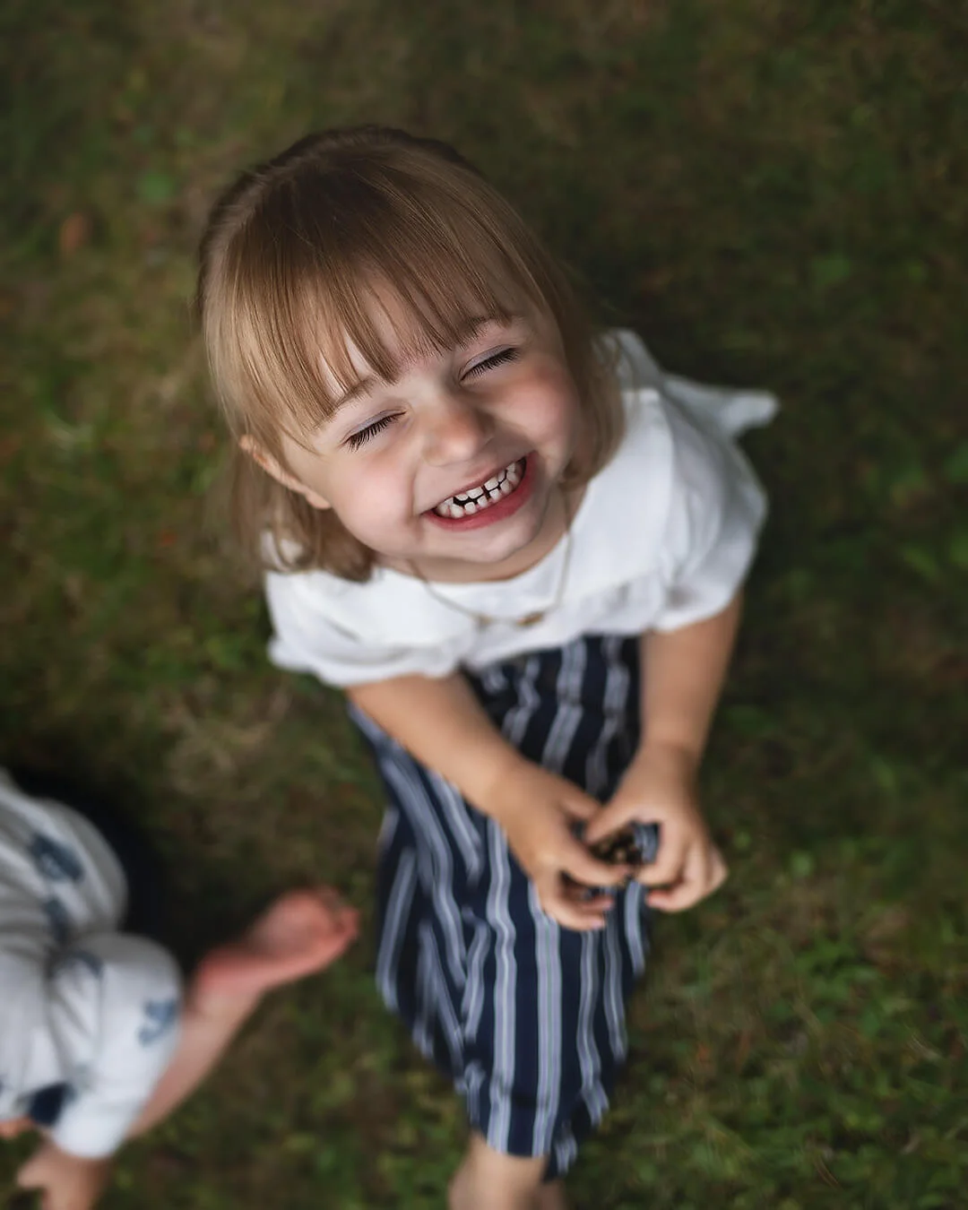 Barnfotografering - porträtt på en flicka sitter på gräset och busar sig framför kameran. Bilden tagen uppifrån. Fotografering i Sala. Sala silvergruva. Children's photography - portrait of a girl sitting on the grass and fooling around in front of the camera. The picture taken from above.