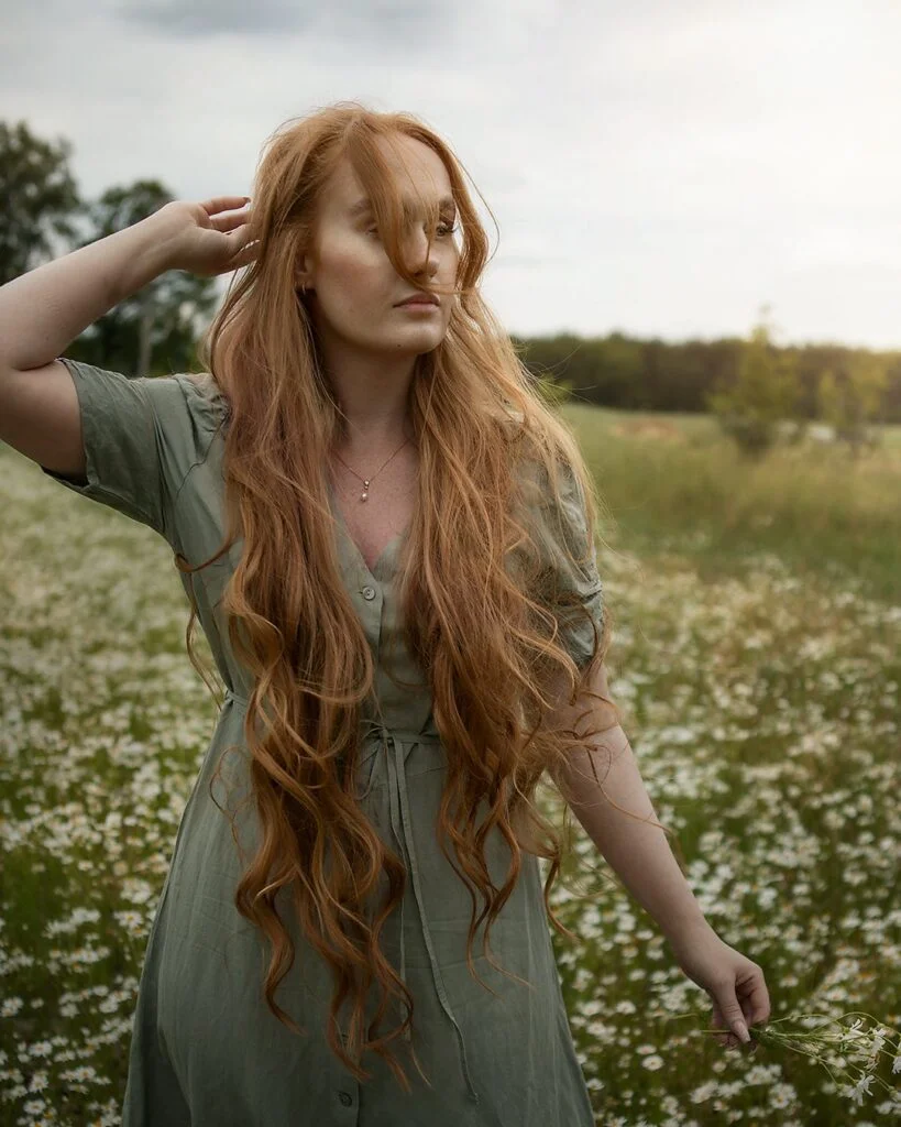 Porträttfotografering - kvinna med rött långt hår går på en äng med vita blommor. Portrait photography - woman with red long hair walking on a meadow with white flowers.