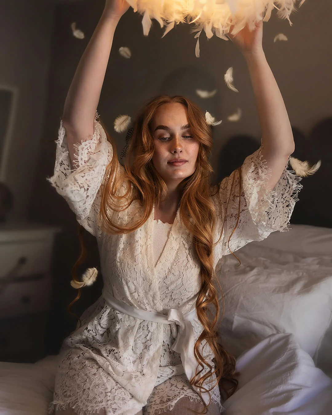 Porträttfotografering - en kvinna (Natali Skoglund) med rött vågigt hår sitter i sängen. Fjädrar faller ner från lampan. Portrait photography - a woman (Natali Skoglund) with red wavy hair is sitting in bed. Feathers fall from the lamp.