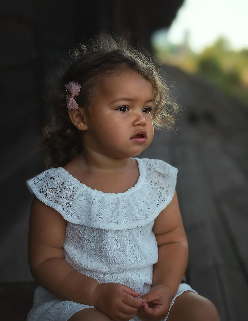 Barnfotografering - en liten flicka med lockigt hår och bruna ögon sitter ned och tittar åt sidan. Barnfoto Sala. Child photography - a little girl with curly hair and brown eyes sits down and looks to the side.