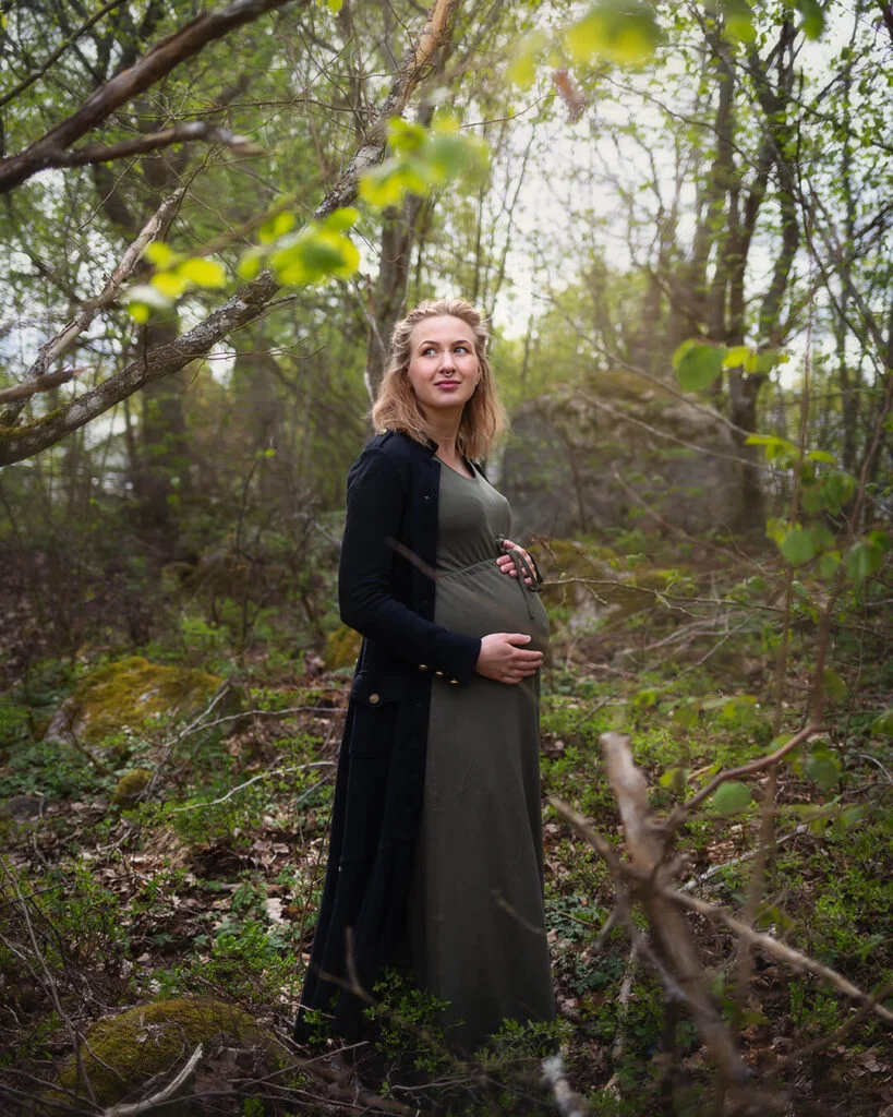 Gravidfotografering - porträtt av en gravid kvinna i grön lång klänning ståendes i skogen och håller sina händer på gravidmagen. gravidfoto i skogen, strängnäs. Maternity photography - portrait of a pregnant woman in a green long dress standing in the forest and holding her hands on her pregnant belly.