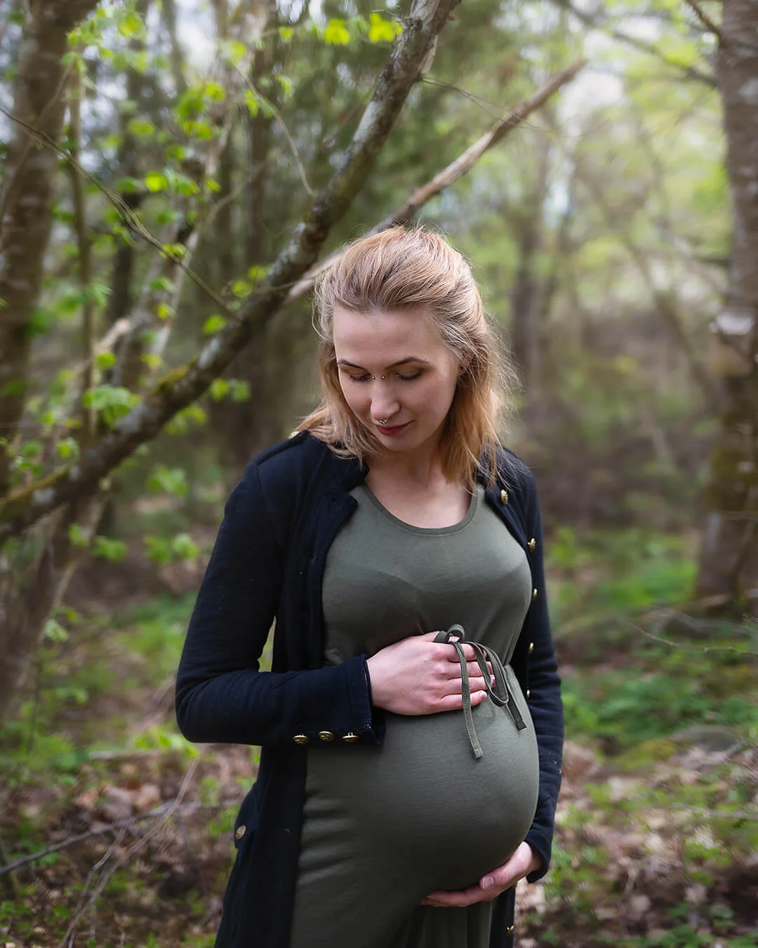 Gravidfotografering - en gravid kvinna står i skogen och håller om sin mage. Gravidfoto i Strängnäs. Maternity photography - a pregnant woman stands in the forest and holds her belly.