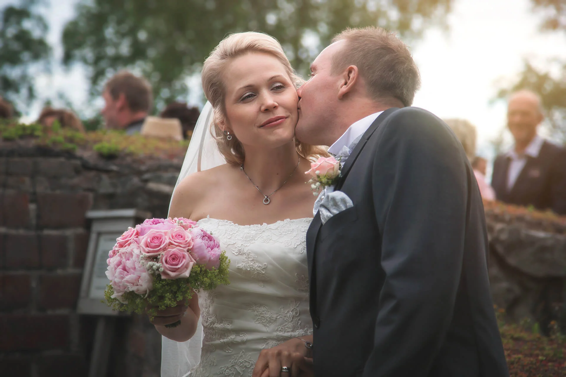 Bröllopsfotografering - brudgum och brud håller hand och brudgummen ger bruden en puss på kinden. Bruden ler. Wedding photography - groom and bride hold hands and the groom gives the bride a kiss on the cheek. The bride smiles.