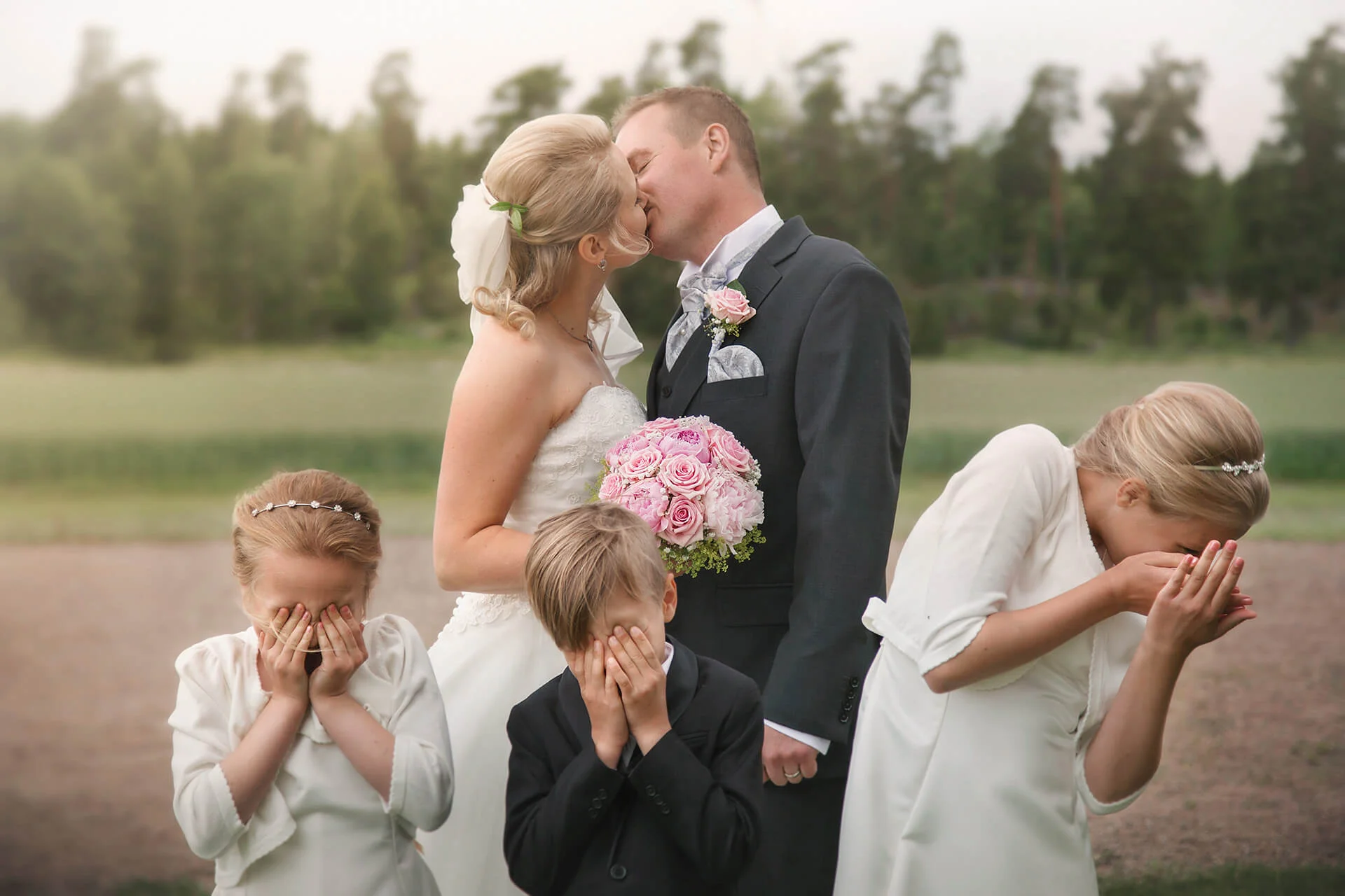Bröllopsfotografering - bröllopsporträtt. brudparet kysser varandra och deras barn (tärnorna och best man) står framför och blundar. Wedding photography - wedding portrait. the bride and groom kiss each other and their children (the bridesmaids and best man) stand in front and close their eyes.