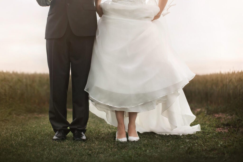 Bröllopsfotografering - bröllops porträtt, brudparet står mot kameran sida vid sida med ett vetefält i bakgrunden, bilden är tagen på underkroppen. Wedding photography - wedding portrait, the bride and groom stand facing the camera side by side with a wheat field in the background, the picture is taken from the lower body.