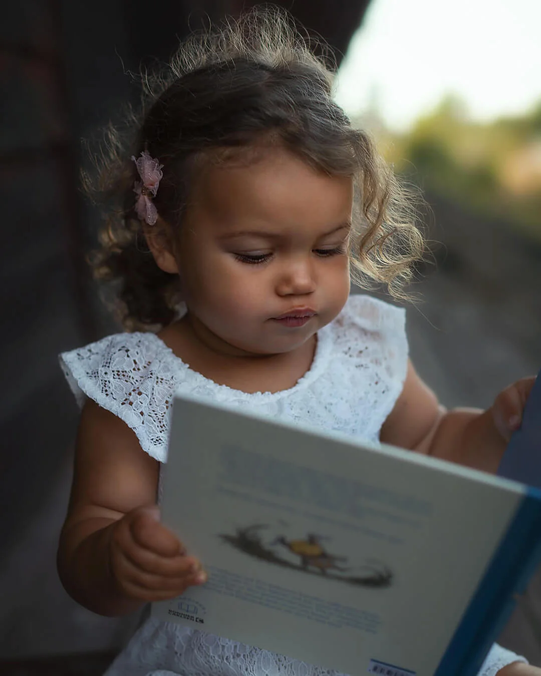 Barnfotografering - en liten flicka med lockigt brunt hår läser en bok. Children's photography - a little girl with curly brown hair is reading a book.