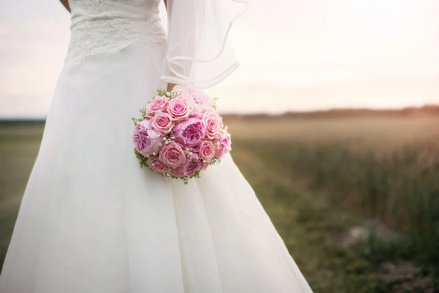 Bröllopsfotografering - bruden står på en åker med rosa brudbukett i fokus. Wedding photography - the bride stands in a field with a pink bridal bouquet in focus.