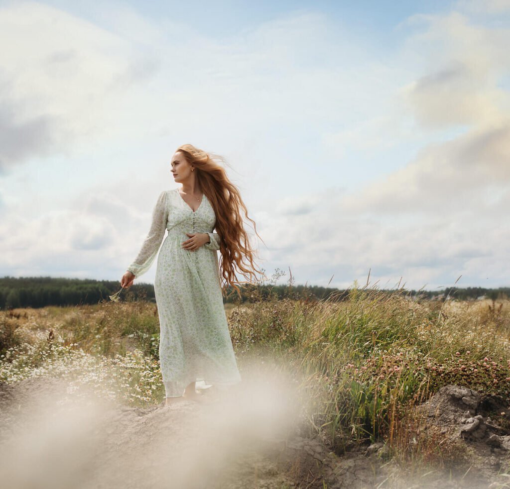 Porträttfotografering - kvinna med långt rött hår klädd i en grön somrig klänning står på en äng. Portrait photography - woman with long red hair wearing a green summer dress stands in a meadow.