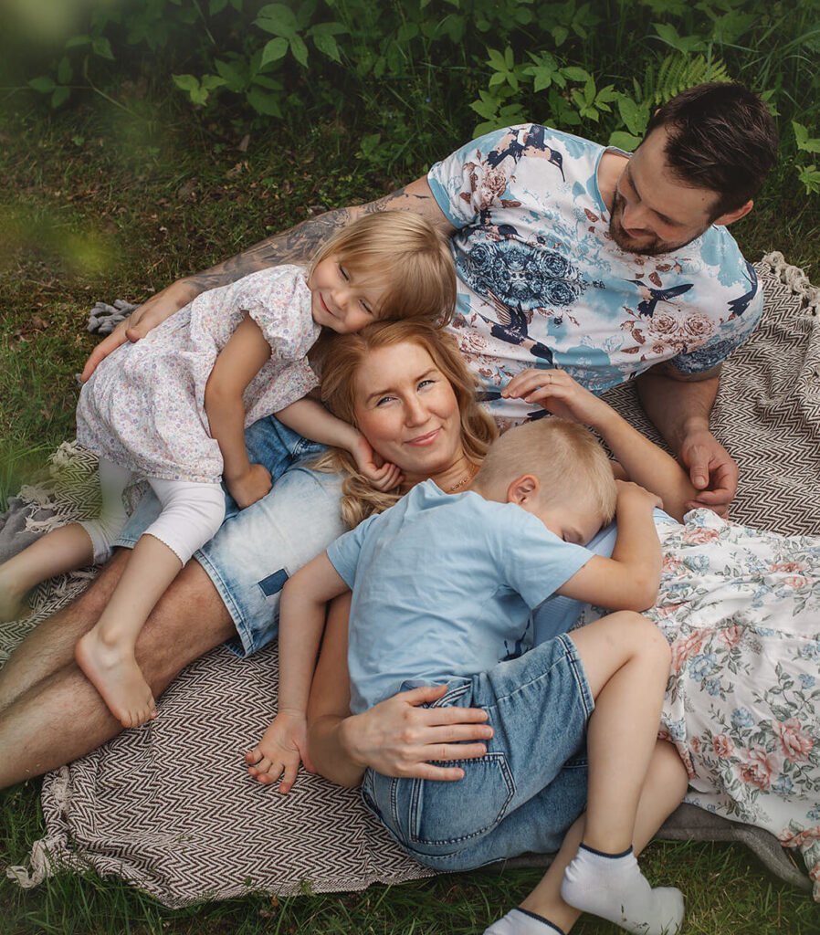 Familjefotografering - Mamma, pappa, och två barn ligger på en filt på gräsmatten och håller om varandra. Family photo shoot - Mom, dad, and two children are lying on a blanket on the lawn and holding each other.