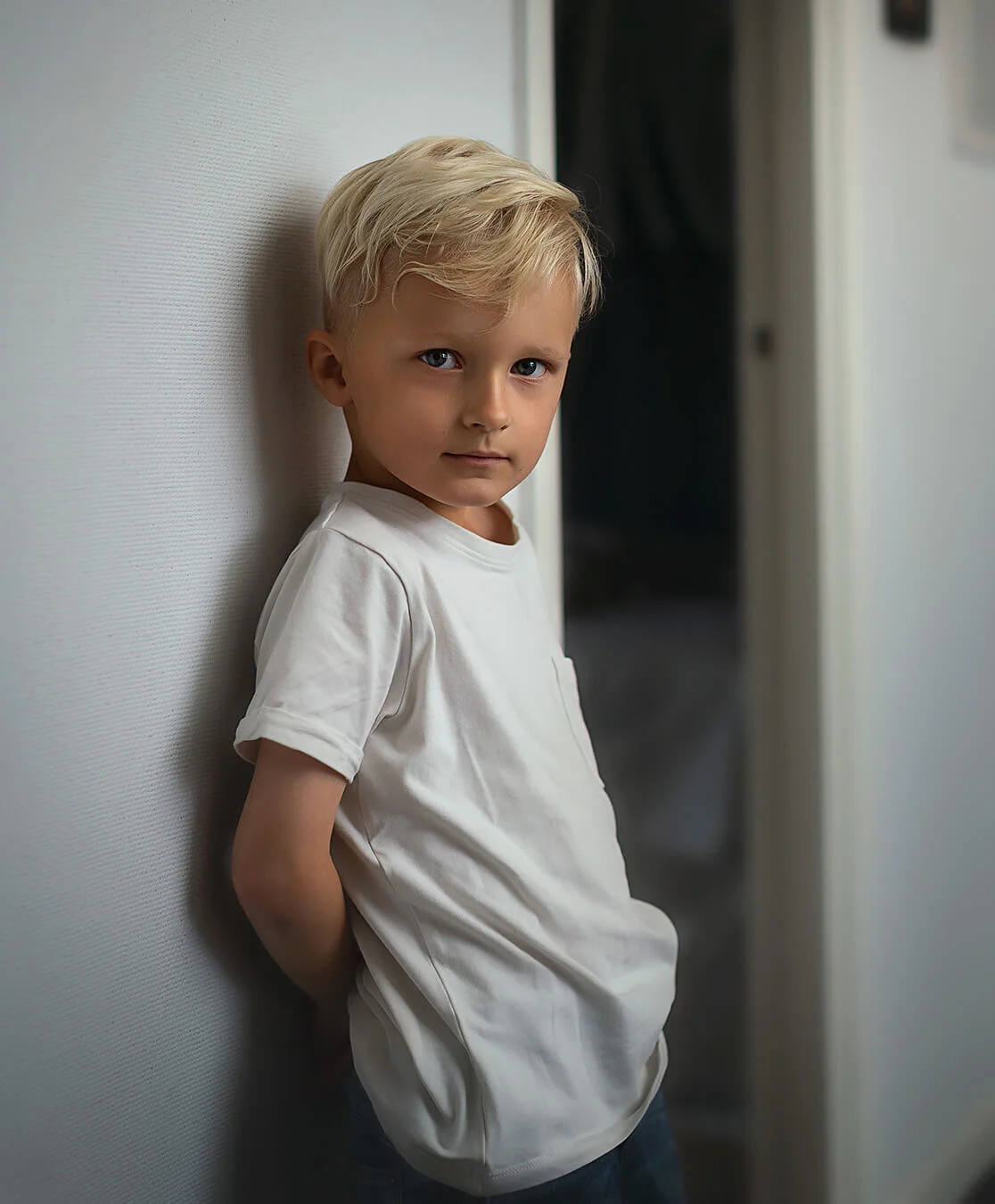 Barnfotografering - porträtt på en pojke som står mot en vid vägg och tittar in i kameran. Children's photography - portrait of a boy standing against a wide wall and looking into the camera.